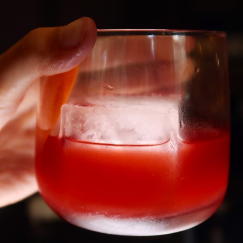 A hand holding a glass with a red cocktail and a large ice cube.