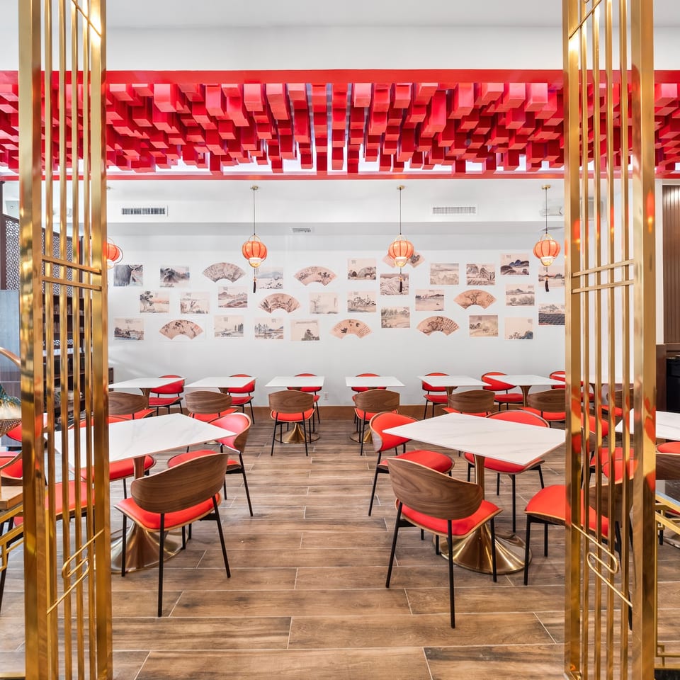 Modern Asian restaurant interior with red chairs, white tables, gold dividers, and a red geometric ceiling.