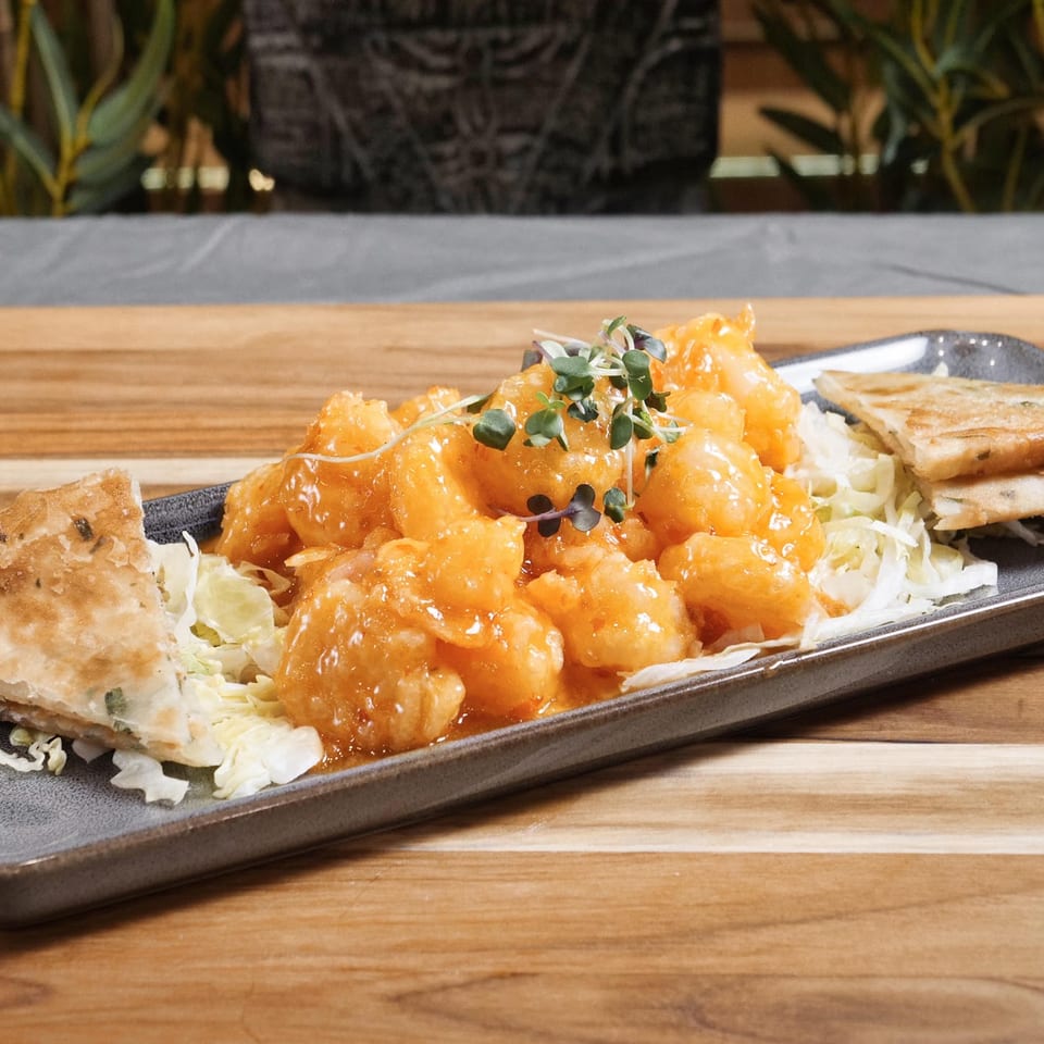 Glazed orange chicken topped with microgreens, shredded cabbage, and crispy flatbread wedges on a rectangular platter.