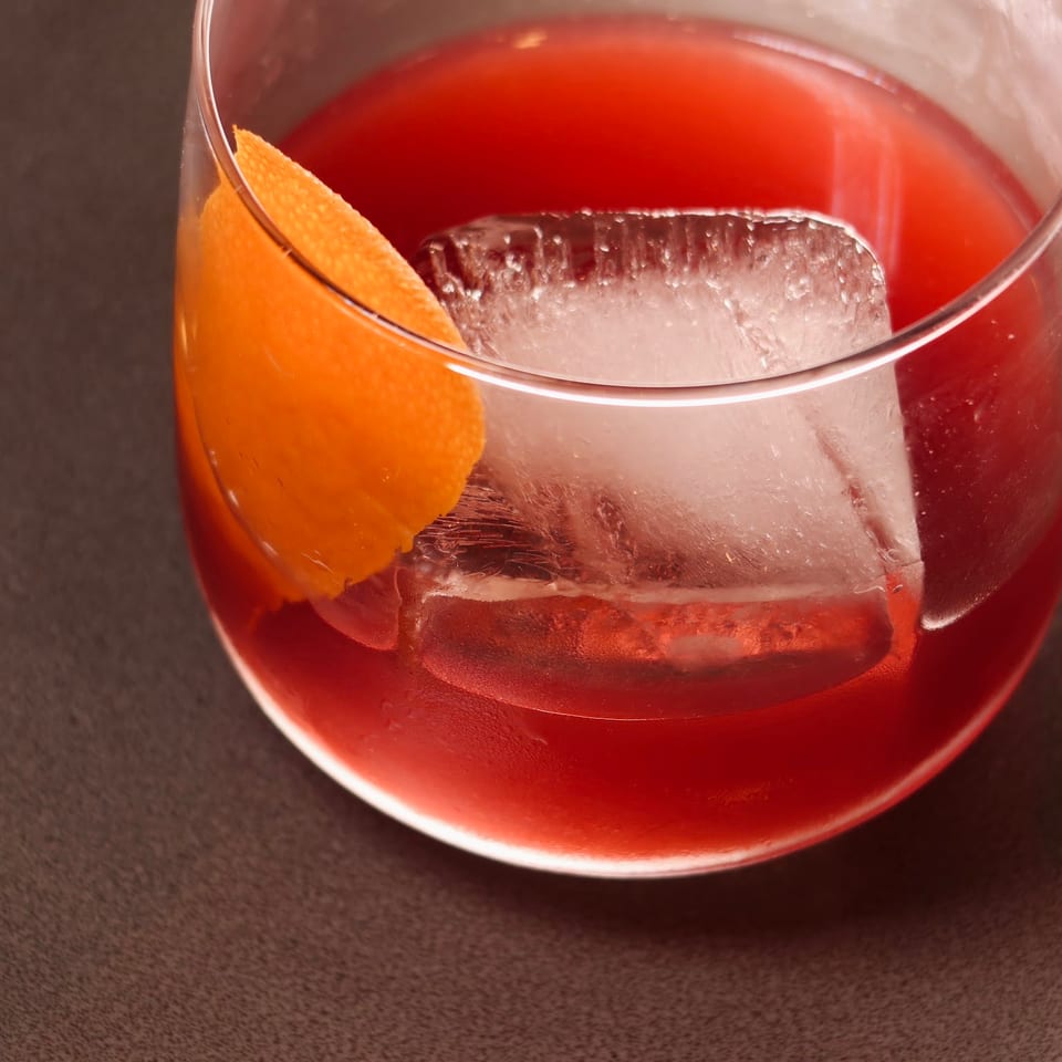 A vibrant red cocktail with a large ice cube and an orange peel garnish in a glass.
