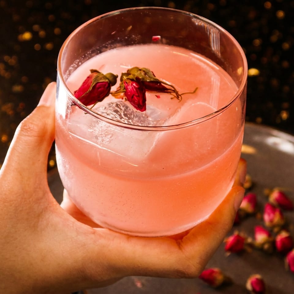 A hand holding a pink cocktail with ice and dried rosebuds, with more rosebuds on a plate.