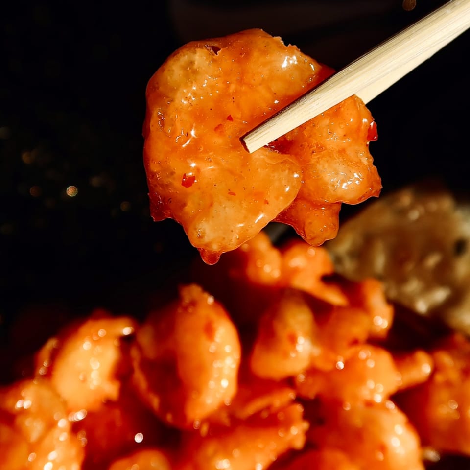 Chopsticks holding a piece of orange-glazed shrimp over a plate of more shrimp.