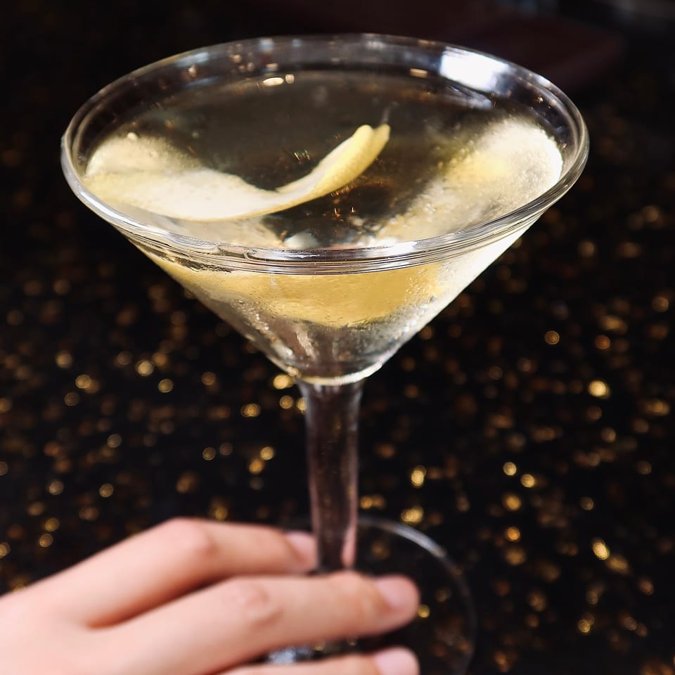 A hand holding a martini glass with a lemon twist against a dark, sparkling background.