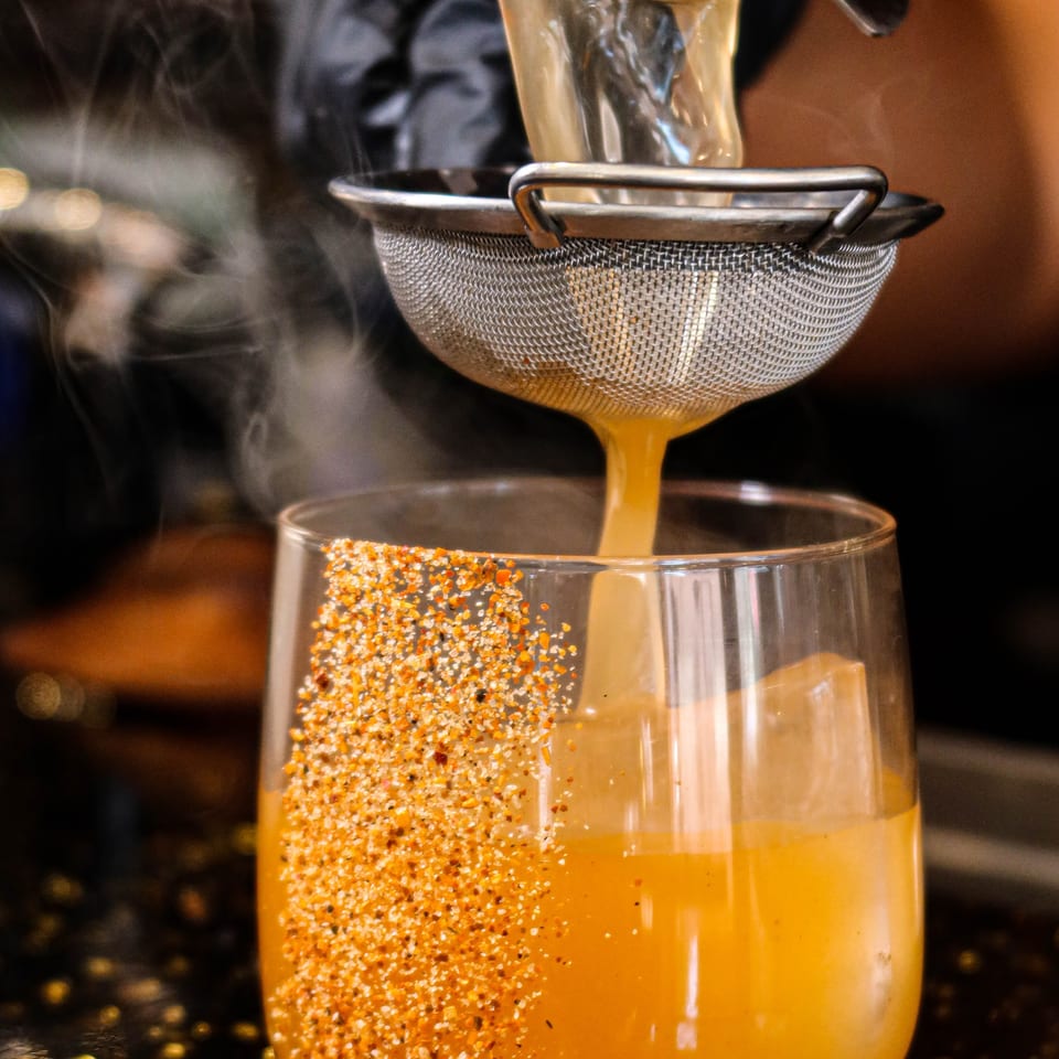 Orange cocktail being strained into a glass rimmed with spices, with steam rising.