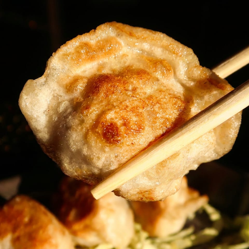 Chopsticks holding a golden-brown fried dumpling, with more dumplings and greens blurred in the background.