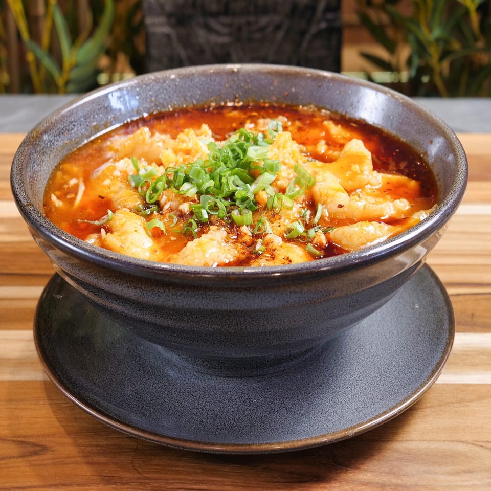 Spicy red broth with dumplings and chopped green onions served in a black ceramic bowl on a wooden table.