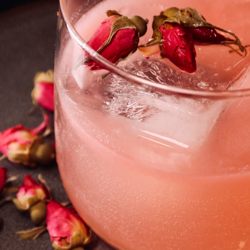 Pink cocktail with ice and two dried rosebuds floating, surrounded by more rosebuds.